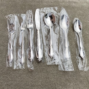 May include: A set of six pieces of silverware, including forks, a knife, and spoons, all wrapped in clear plastic. The silverware has a shiny, silver finish and a decorative handle design. The set is likely for dining.