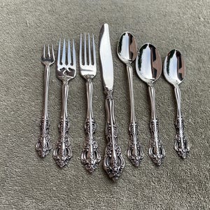 May include: A set of silver-colored flatware, including forks, a knife, and spoons, arranged on a textured surface. The utensils have ornate, detailed handles. The set includes a small fork, two larger forks, a knife, and three spoons.