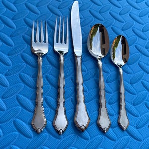 May include: A set of five silver-colored utensils: two forks, a knife, and two spoons. The utensils have ornate handles and are arranged on a textured blue surface. The knife is in the center, flanked by the forks and spoons.