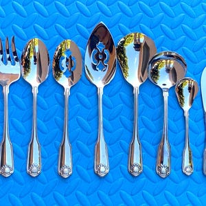 May include: A collection of polished silver serving utensils, including a fork, spoons, and a knife, arranged on a textured blue surface. The utensils have decorative handles and various serving heads, reflecting the light. Ideal for formal dining.
