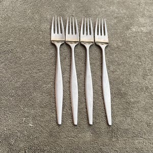May include: Four silver forks with a simple, modern design are arranged vertically on a textured, gray surface. Each fork has a slender handle and four tines. The forks are made of a shiny metal.