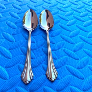 May include: Two polished silver spoons with a decorative handle design. The spoons are oval-shaped and reflect the surrounding environment. The spoons are set against a blue textured background. The spoons are likely for eating.