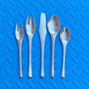 Könnte beinhalten: Ein Set aus fünf silberfarbenen Essbestecken, darunter zwei Gabeln, zwei Löffel und ein Messer, angeordnet vor einem strukturierten blauen Hintergrund. Die Utensilien haben lange, schlanke Griffe und sind wahrscheinlich zum Essen.
