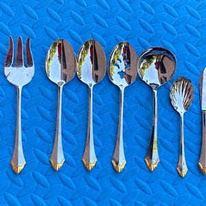 Könnte beinhalten: Ein Set aus sechs silberfarbenen Servierbesteckteilen aus Edelstahl mit goldfarbenen Akzenten am Griffende. Die Kollektion umfasst eine Gabel mit drei Zinken, zwei volle Löffel, einen Schlitzlöffel, eine Schöpfkelle, einen kleinen muschelförmigen Löffel und ein Messer.