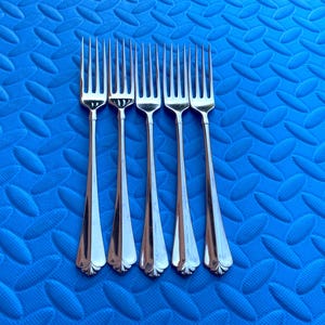 May include: Five silver forks with a classic design are arranged vertically against a textured blue background. The forks have long, slender handles and tines. The background has a repeating pattern of oval shapes.