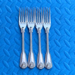 May include: Four silver-colored metal forks arranged side by side. Each fork features a four-tined head and a handle decorated with a raised shell-like motif at the base. The metal has a polished, reflective finish throughout the entire length.