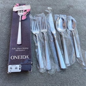 Könnte beinhalten: Ein 5-teiliges Besteckset aus Edelstahl von Oneida, einschließlich Gabeln, Messern und Löffeln. Das Set ist in Kunststoff und einer dunkelvioletten Schachtel mit Markennamen und Produktdetails verpackt. Die Gabel in der Schachtel ist sichtbar.
