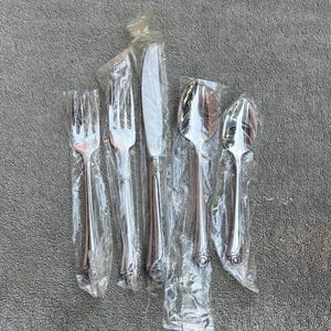 May include: Five pieces of cutlery, including a knife, two forks, and two spoons, are wrapped in plastic. The cutlery is silver in colour and has ornate designs.