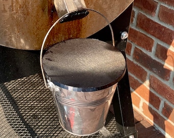 Op de afbeelding: Een zilveren metalen emmer met een zwarte deksel staat op een zwarte metalen standaard. De emmer hangt aan een metalen haak die aan een grote metalen trommel is bevestigd.