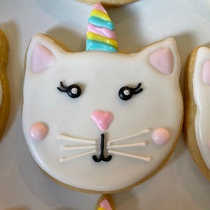 Puede incluir: Una galleta de azúcar blanca decorada con glaseado rosa y blanco para parecer un gato con un cuerno de unicornio arcoíris.
