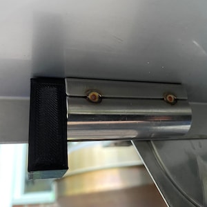 May include: A close-up of a black metal bracket attached to a silver metal pipe. The bracket has two small, round, gold-colored screws.