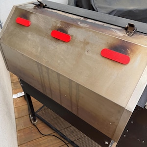 May include: A stainless steel grill with three red handles. The grill has a black lid and is on a black metal frame with a lower shelf. The grill is outdoors on a wooden deck.