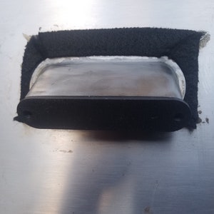 May include: A close-up of a black rectangular metal plate with two screws on the side. The plate is mounted on a silver surface and is surrounded by black foam.
