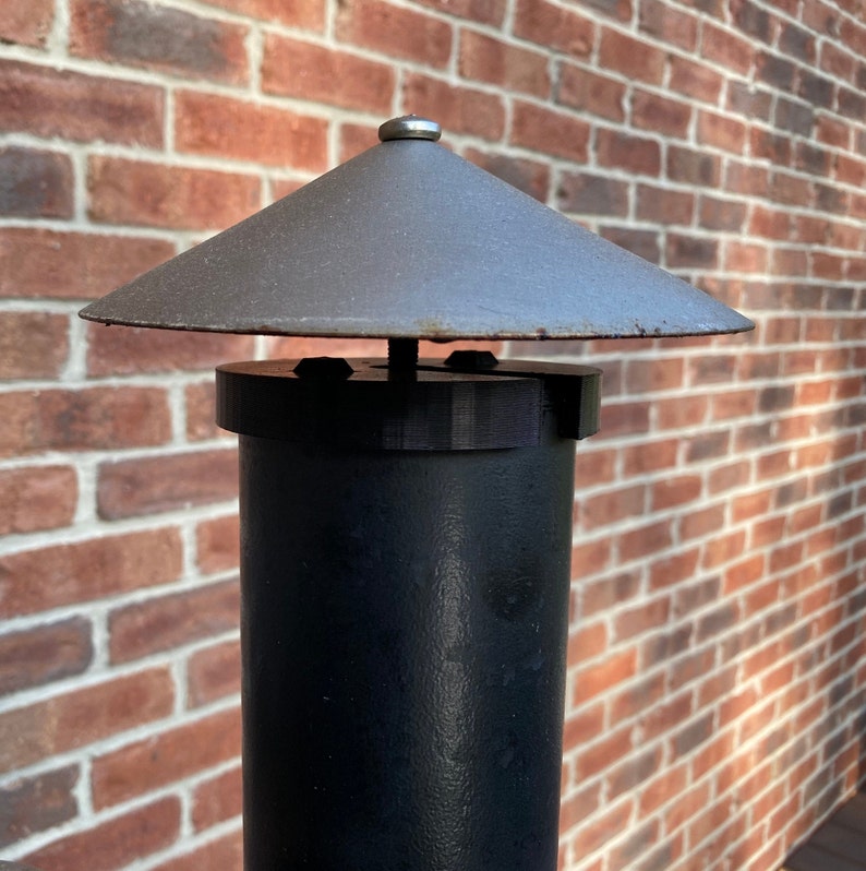Op de afbeelding: Een zwarte metalen paal met een grijze metalen dop. De dop heeft een afgeronde bovenkant en een platte basis. De paal staat tegen een bakstenen muur.
