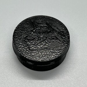 May include: A black round container with a textured surface and a raised design of a stylized animal head.