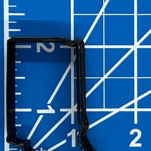 以下が含まれることがあります： インディアナ州の形をした黒い金属製のクッキーカッター。クッキーカッターの高さは約5cm、幅は約2.5cmです。白いグリッドラインのある青いカッティングマットの上に置かれています。