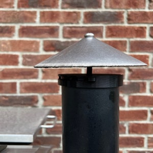 Puede incluir: Una chimenea de metal negro con una parte superior cónica gris. La tapa está montada en una tubería cilíndrica negra. El fondo presenta una pared de ladrillo rojo.