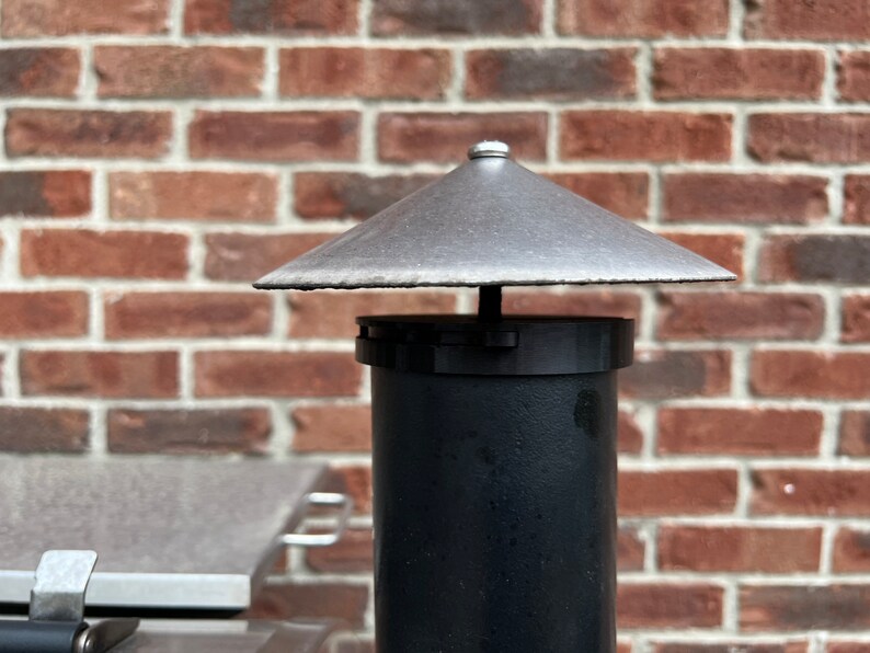 May include: A black metal chimney pipe with a silver metal rain cap. The pipe is against a brick wall.