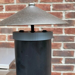Op de afbeelding: Een zwarte metalen schoorsteenpijp met een grijze metalen kap en een grijze metalen regenkap. De schoorsteenpijp is bevestigd aan een bakstenen muur.
