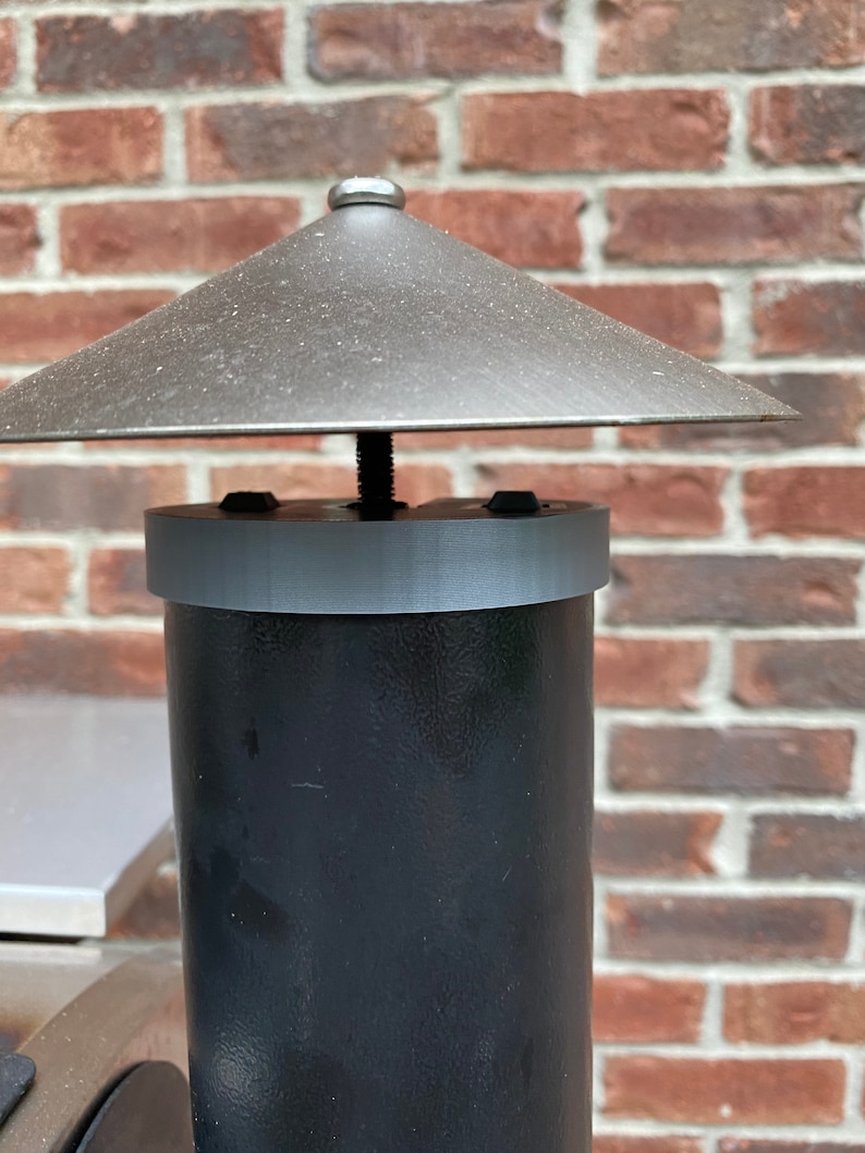 Op de afbeelding: Een zwarte metalen schoorsteenpijp met een grijze metalen kap en een grijze metalen regenkap. De schoorsteenpijp is bevestigd aan een bakstenen muur.