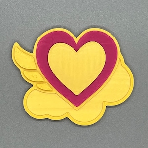 Puede incluir: Un corazón amarillo impreso en 3D con un corazón rosa en el interior, con alas a los lados.