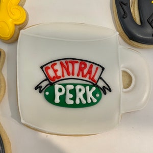 May include: A white coffee mug shaped sugar biscuit decorated with royal icing. The centre features a red and black banner with the text CENTRAL and a green oval below with the white text PERK. The biscuit has a handle on the right side.
