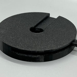 May include: A black plastic spool with a small, removable tab on the side. The spool has a central hole and a groove around the edge.