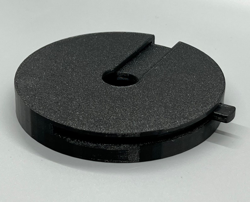 May include: A black plastic spool with a small, removable tab on the side. The spool has a central hole and a groove around the edge.