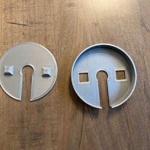 Op de afbeelding: Twee grijze plastic cirkels met een sleutelgat uitsnijding. E&eacute;n cirkel heeft twee vierkante uitsnijdingen aan de zijkant, de andere heeft twee vierkante bulten aan de zijkant.
