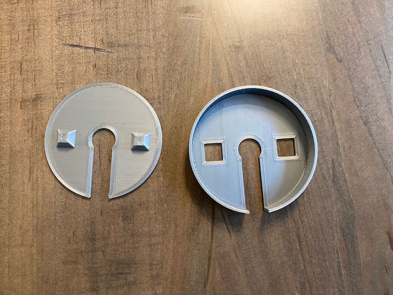 Op de afbeelding: Twee grijze plastic cirkels met een sleutelgat uitsnijding. E&eacute;n cirkel heeft twee vierkante uitsnijdingen aan de zijkant, de andere heeft twee vierkante bulten aan de zijkant.