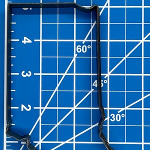 以下が含まれることがあります： インディアナ州の形をした黒い金属製のクッキーカッター。カッターの高さは7.6cm、幅は5cmです。青いカッティングマットに白いグリッドラインが引かれた状態で表示されています。