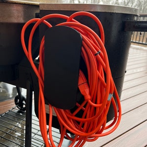 May include: A black metal extension cord holder attached to a black grill. An orange extension cord is wrapped around the holder.
