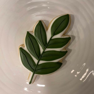 May include: A green leaf-shaped cookie decorated with white icing. The cookie is on a white plate.