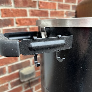 Könnte beinhalten: Nahaufnahme eines schwarzen Metall-Smokers mit einem silbernen Deckel und zwei Haken an der Seite.
