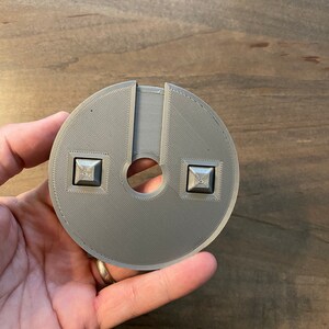Op de afbeelding: Een grijs 3D-geprint cirkelvormig object met twee vierkante knoppen en een gat in het midden.