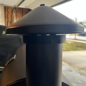 May include: Close-up of a black metal chimney cap for a smoker or grill. The cap has a round, cone-shaped top and a cylindrical base.