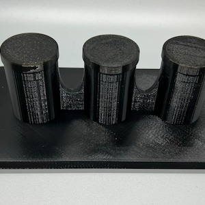 Pode incluir: Um suporte preto impresso em 3D com tr&ecirc;s suportes cil&iacute;ndricos. O suporte &eacute; projetado para segurar itens pequenos, como canetas, l&aacute;pis ou pinc&eacute;is.