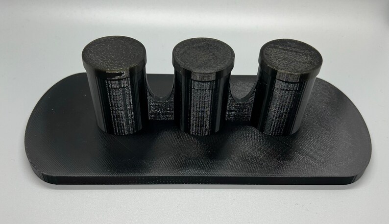 Pode incluir: Um suporte preto impresso em 3D com tr&ecirc;s suportes cil&iacute;ndricos. O suporte &eacute; projetado para segurar itens pequenos, como canetas, l&aacute;pis ou pinc&eacute;is.