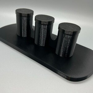 Pode incluir: Um suporte preto impresso em 3D com tr&ecirc;s suportes cil&iacute;ndricos. O suporte &eacute; projetado para segurar itens pequenos como canetas, l&aacute;pis ou pinc&eacute;is.