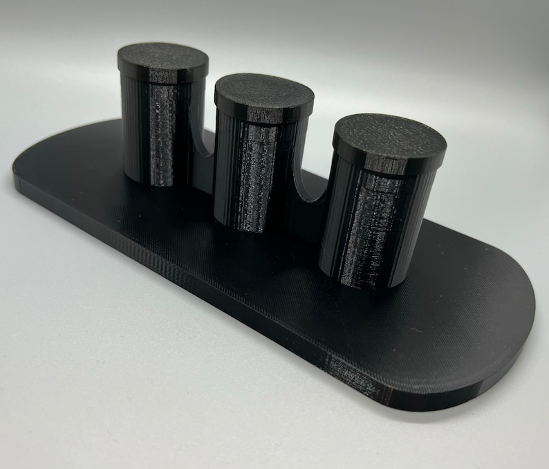 Pode incluir: Um suporte preto impresso em 3D com tr&ecirc;s suportes cil&iacute;ndricos. O suporte &eacute; projetado para segurar itens pequenos como canetas, l&aacute;pis ou pinc&eacute;is.