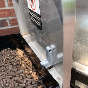 Puede incluir: Primer plano de una tolva de pellets para barbacoa llena de pellets de madera. La tolva tiene una tapa de metal con una etiqueta de advertencia que dice "PELIGRO - SIN FIN ROTATORIO - Nunca introduzca las manos u objetos en la tolva mientras el sin fin esté en funcionamiento. Puede producirse una lesión grave."