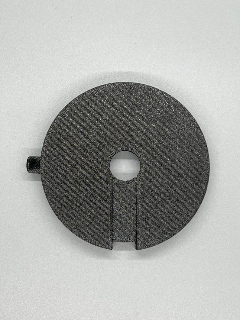 May include: A black plastic 3D printed adapter with a round shape and a hole in the centre. The adapter has a small tab on the side.