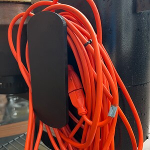 Pode incluir: Um suporte de cabo de pl&aacute;stico preto com um cabo de extens&atilde;o laranja enrolado ao redor.