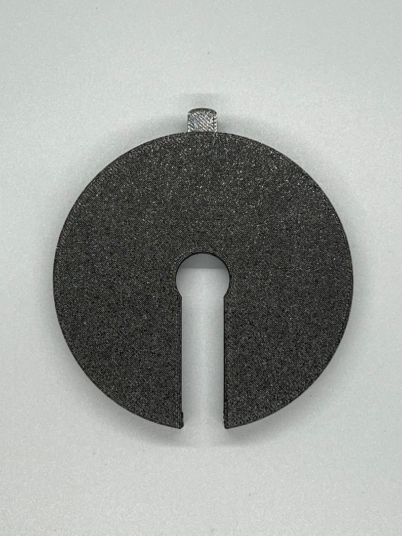 May include: A black plastic disc with a keyhole-shaped cutout. The disc has a textured surface and a small loop at the top.