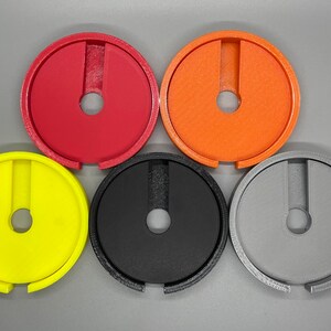 Op de afbeelding: Vijf plastic ringen in verschillende kleuren: rood, oranje, geel, zwart en grijs. Elke ring heeft een kleine opening in het midden en een grotere opening aan de zijkant.