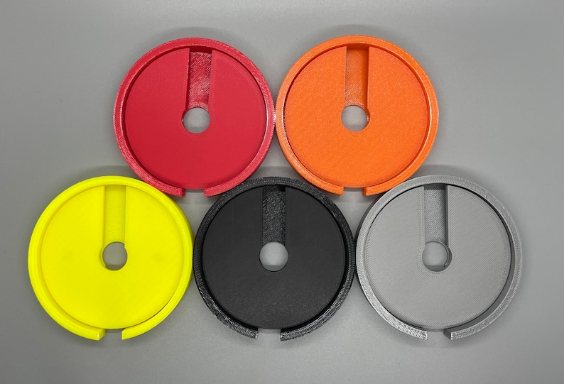 Op de afbeelding: Vijf plastic ringen in verschillende kleuren: rood, oranje, geel, zwart en grijs. Elke ring heeft een kleine opening in het midden en een grotere opening aan de zijkant.