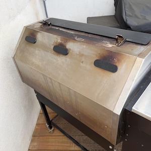 May include: A stainless steel grill with a black lid and accents. The grill has a rectangular shape with a black metal frame and a lower shelf. The grill is on wheels and appears to be used.