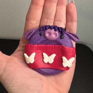 May include: A purple, handmade clay figurine shaped like a pig with a red band featuring three white butterfly cutouts. The pig has a pink snout, black eyes, and purple hair.