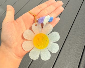 Peut inclure: Un porte-clés en forme de marguerite avec un centre jaune en forme de smiley et des pétales blancs. Le porte-clés a un anneau doré et un pompon avec des brins violets, blancs et bleus. Le fond est une surface grise.
