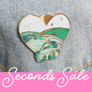 May include: A gold heart-shaped enamel pin with a white background and a green and blue landscape design. The pin features a stylised depiction of rolling hills and clouds.